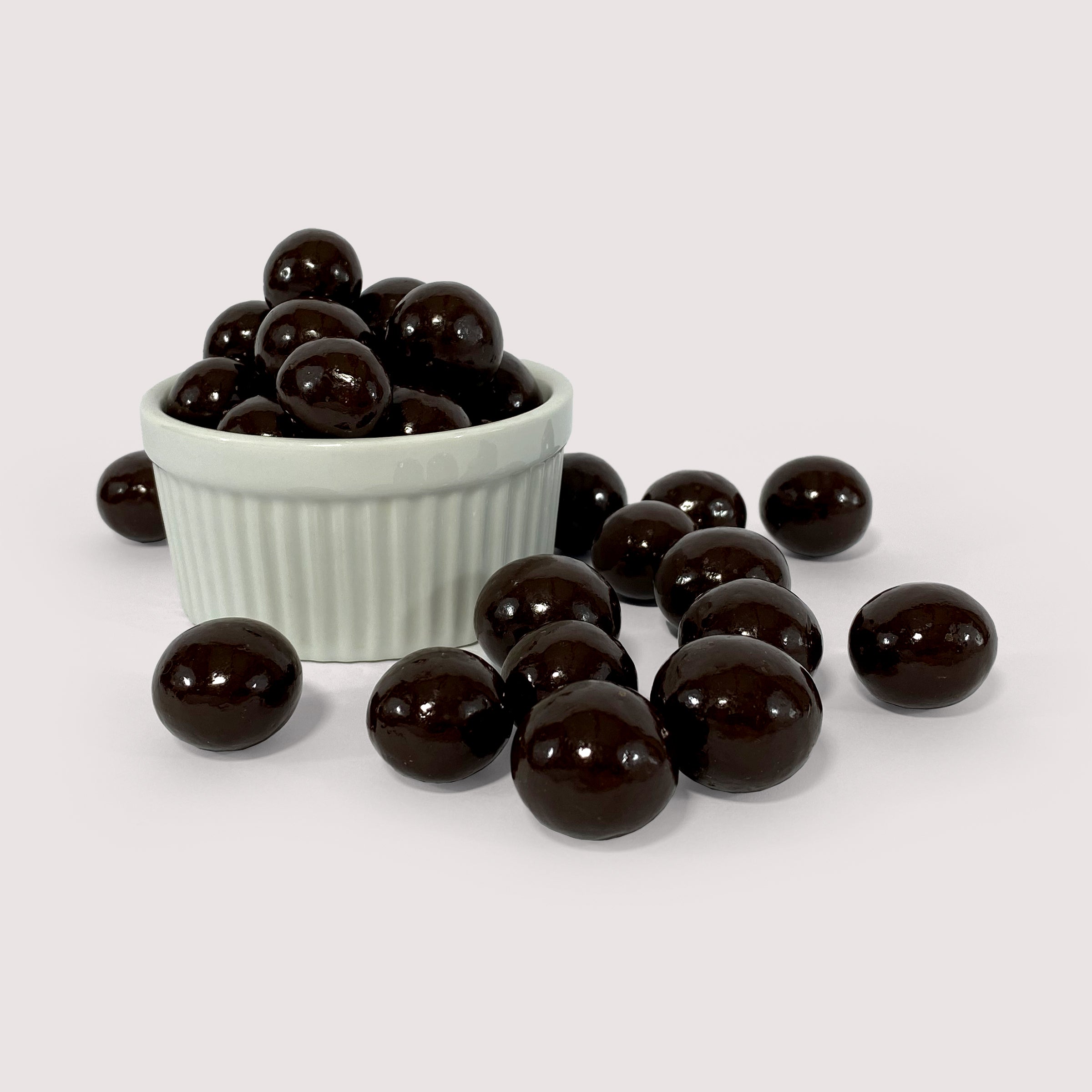 Image shows a pile of dark chocolate coated macadamia nuts displayed in a fluted white bowl with some dark chocolate coated macadamia nuts scattered in front 