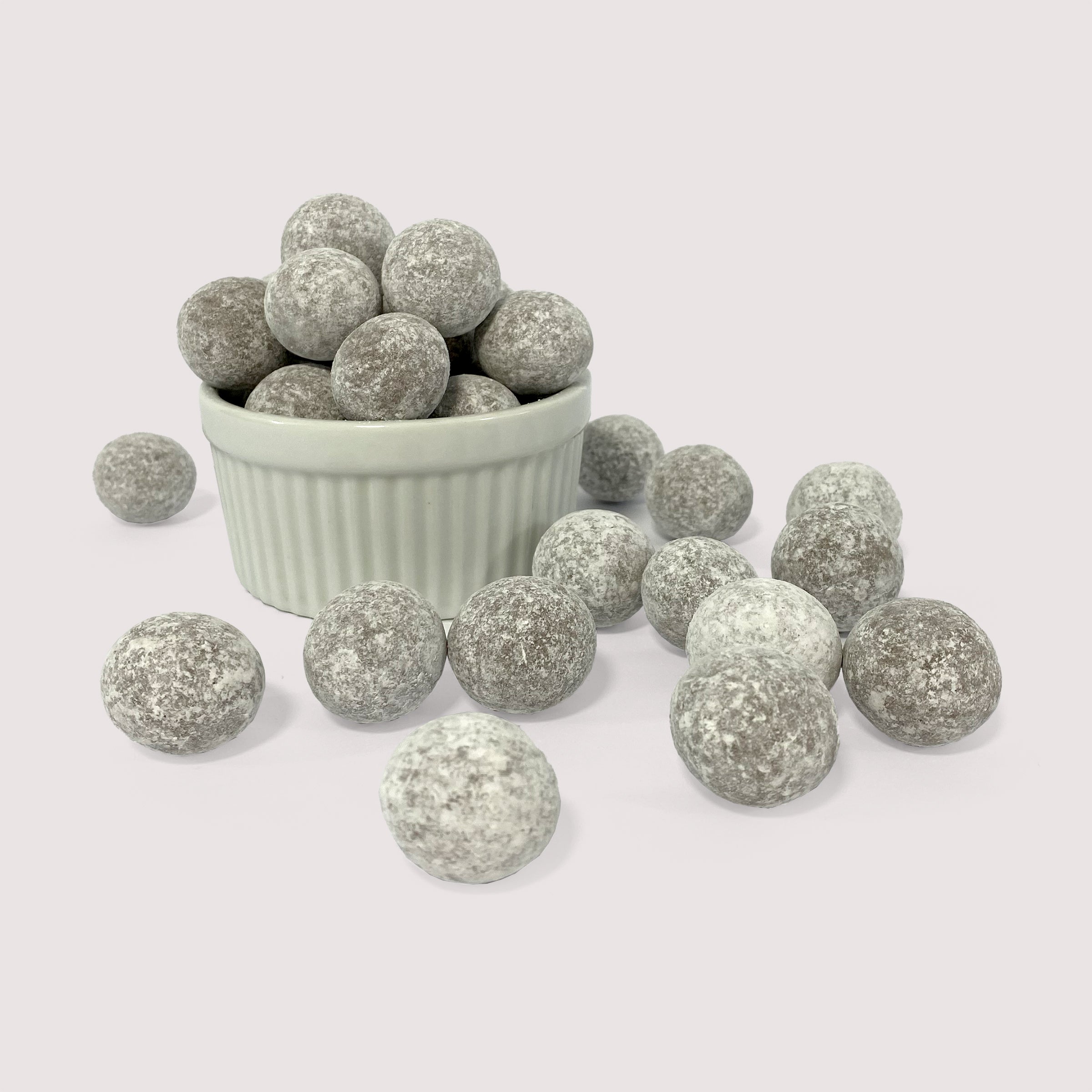 Image shows a pile of powdered sugar coated chocolate coconut flavored toffee macadamia nuts displayed in a fluted white bowl with some powdered sugar coated chocolate coconut flavored toffee macadamia nuts scattered in front 