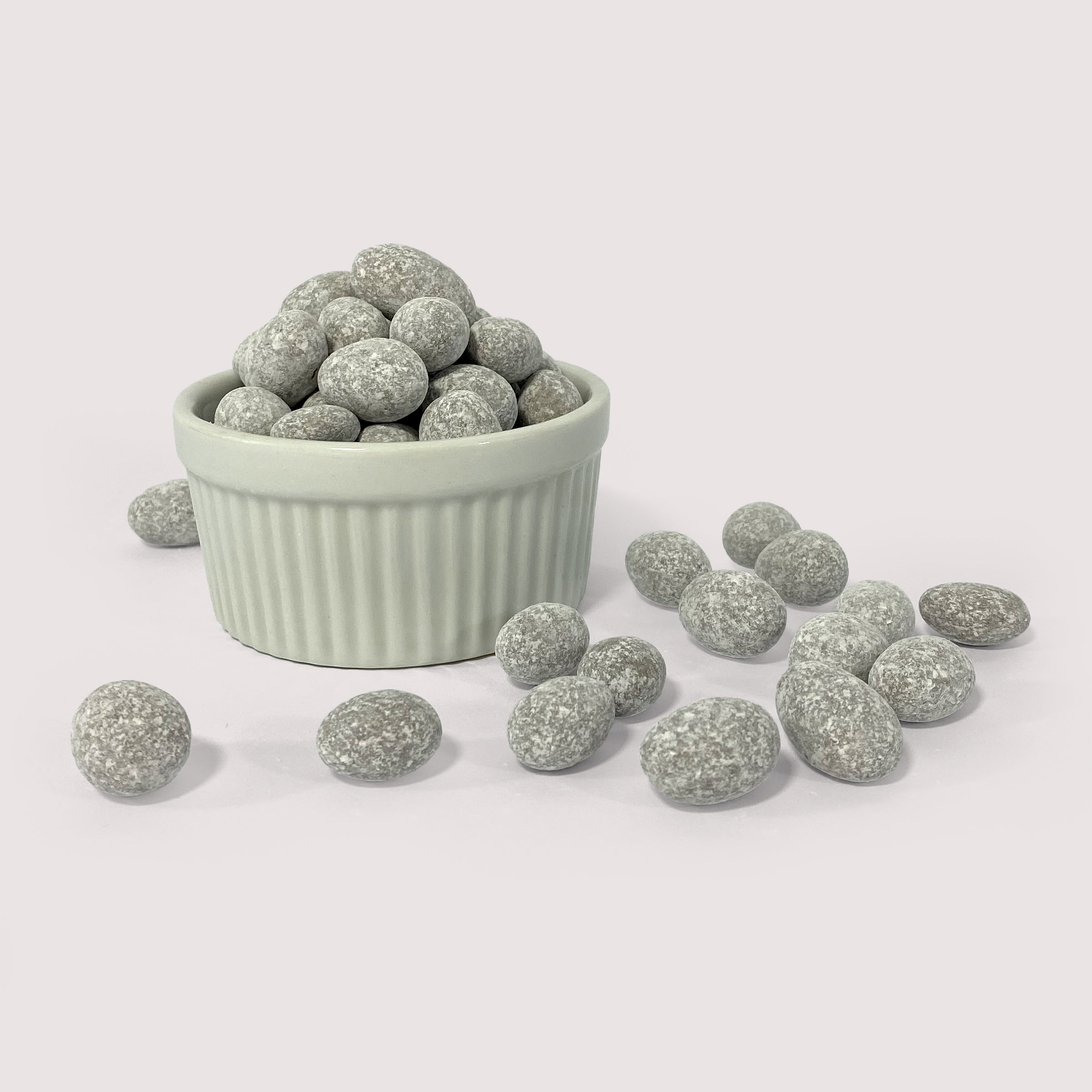 Image shows a pile of powdered sugar coated chocolate toffee almonds displayed in a fluted white bowl with some powdered sugar coated chocolate toffee almonds scattered in front 