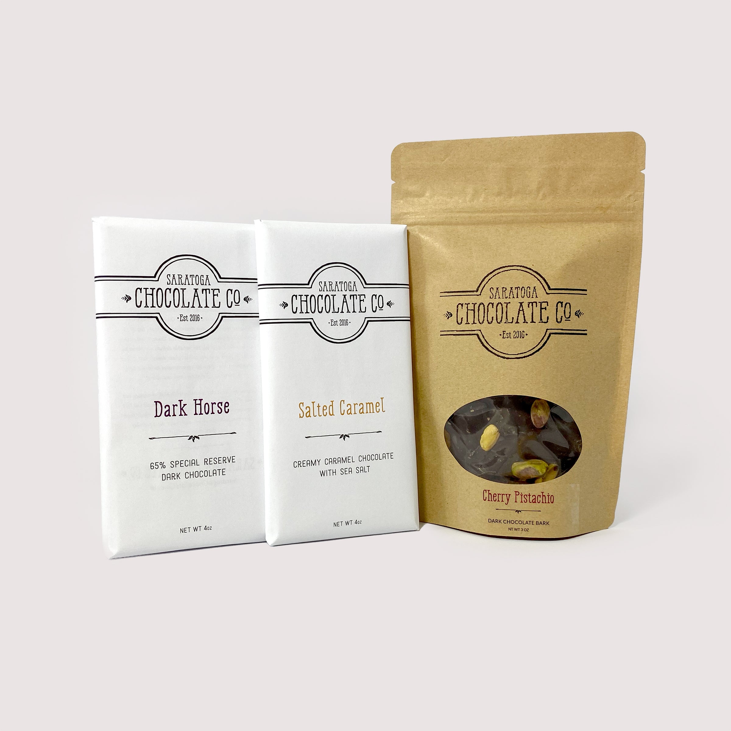 Saratoga Chocolate Co. '2 Bars and a Bark' set featuring two 4-ounce chocolate bars in white wrappers with flavor labels, alongside a 3-ounce resealable kraft brown bag of chocolate bark with a clear viewing window displaying the bark's inclusions.