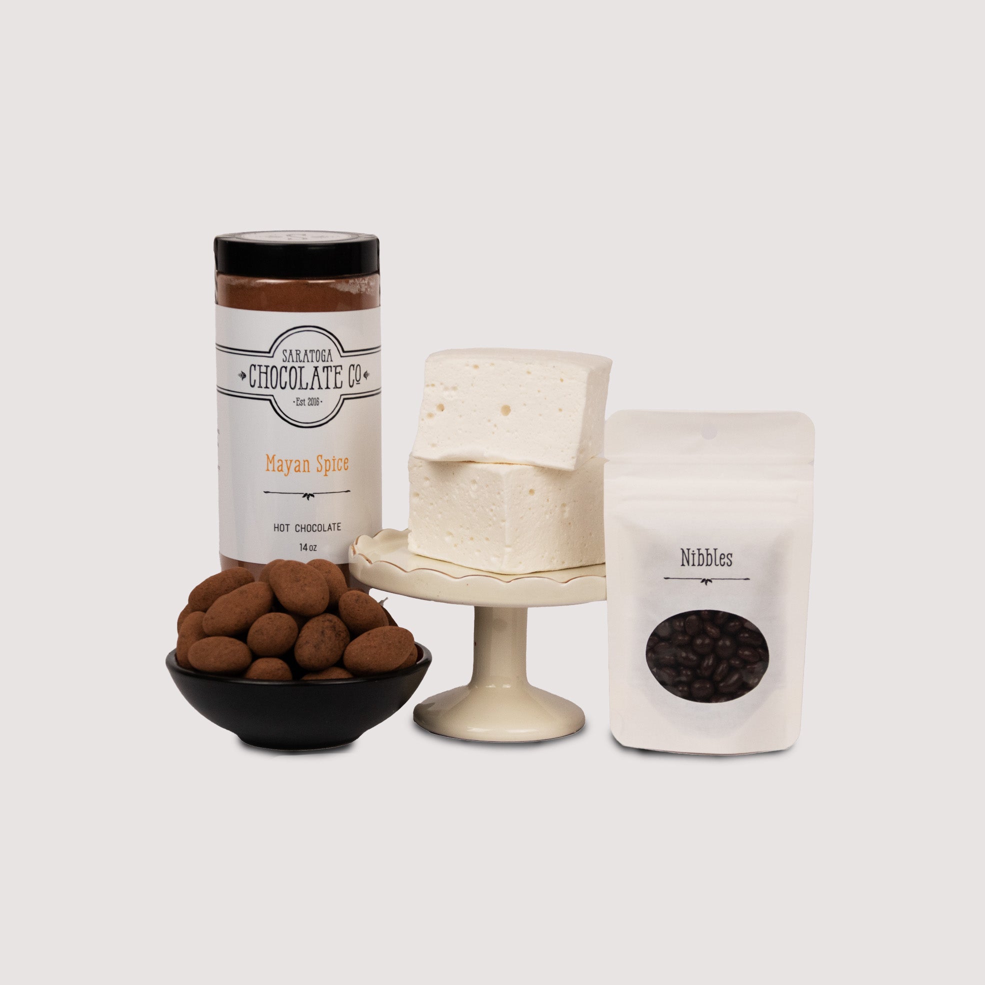 This image shows a cluster of products that make up the "Spicy Date" gift set. The items shown include a 14 ounce jar of Mayan Spice hot chocolate mix, two pillow-soft, snow white vanilla marshmallows, a 1.5oz pouch of chocolate covered cacao nib "Nibbles", and a pile of cocoa powder dusted Mexican Vanilla chocolate covered Almonds.