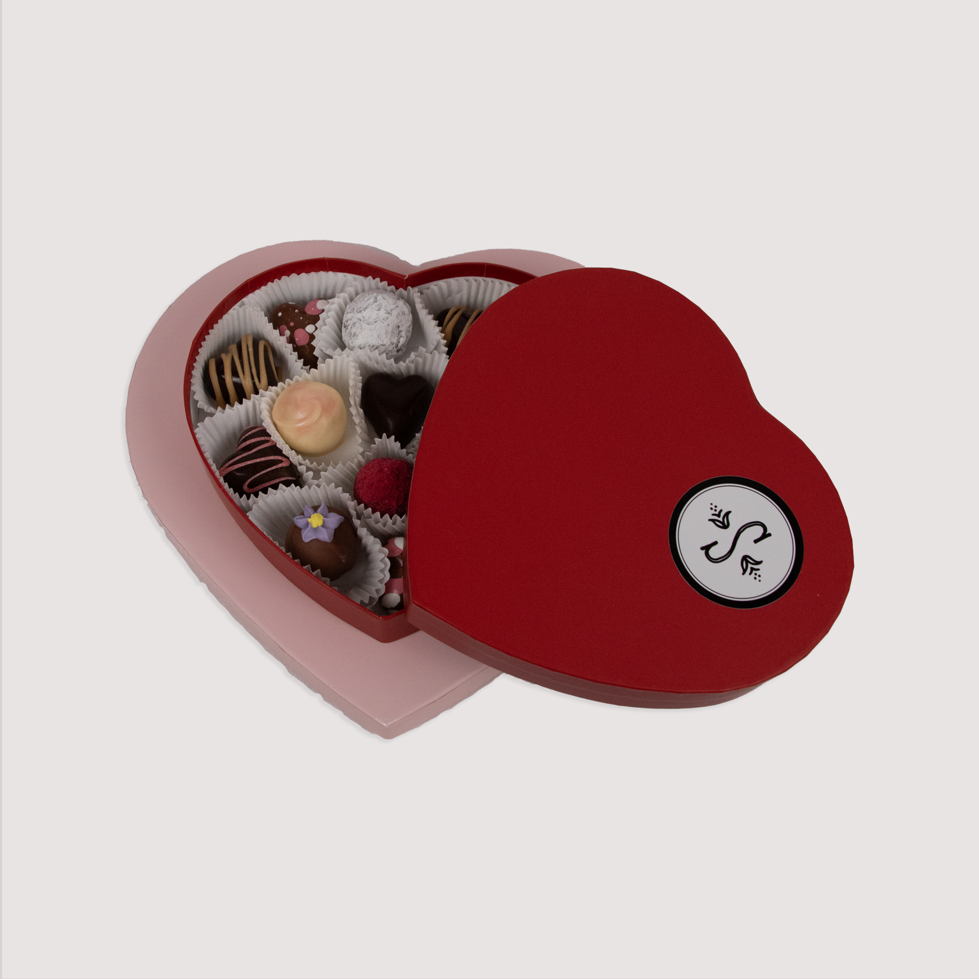 A red heart-shaped box with Saratoga Chocolate logo filled with assorted chocolate truffles in various flavors and designs, arranged neatly in individual paper cups. 