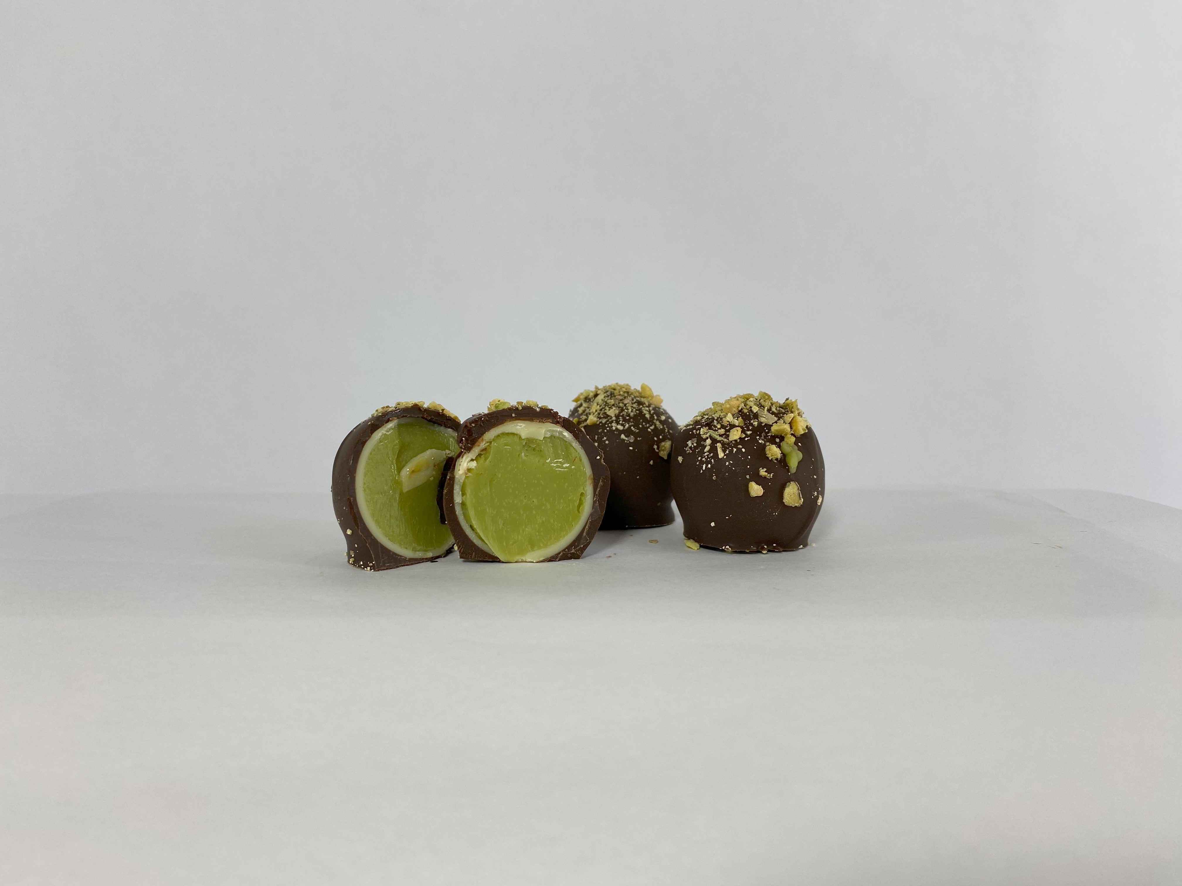Close-up of a Saratoga Chocolate Co. Pistachio Truffle sliced in half, revealing a smooth, nutty pistachio ganache encased in a white chocolate layer, all enveloped by a dark chocolate shell and garnished with ground salted pistachios