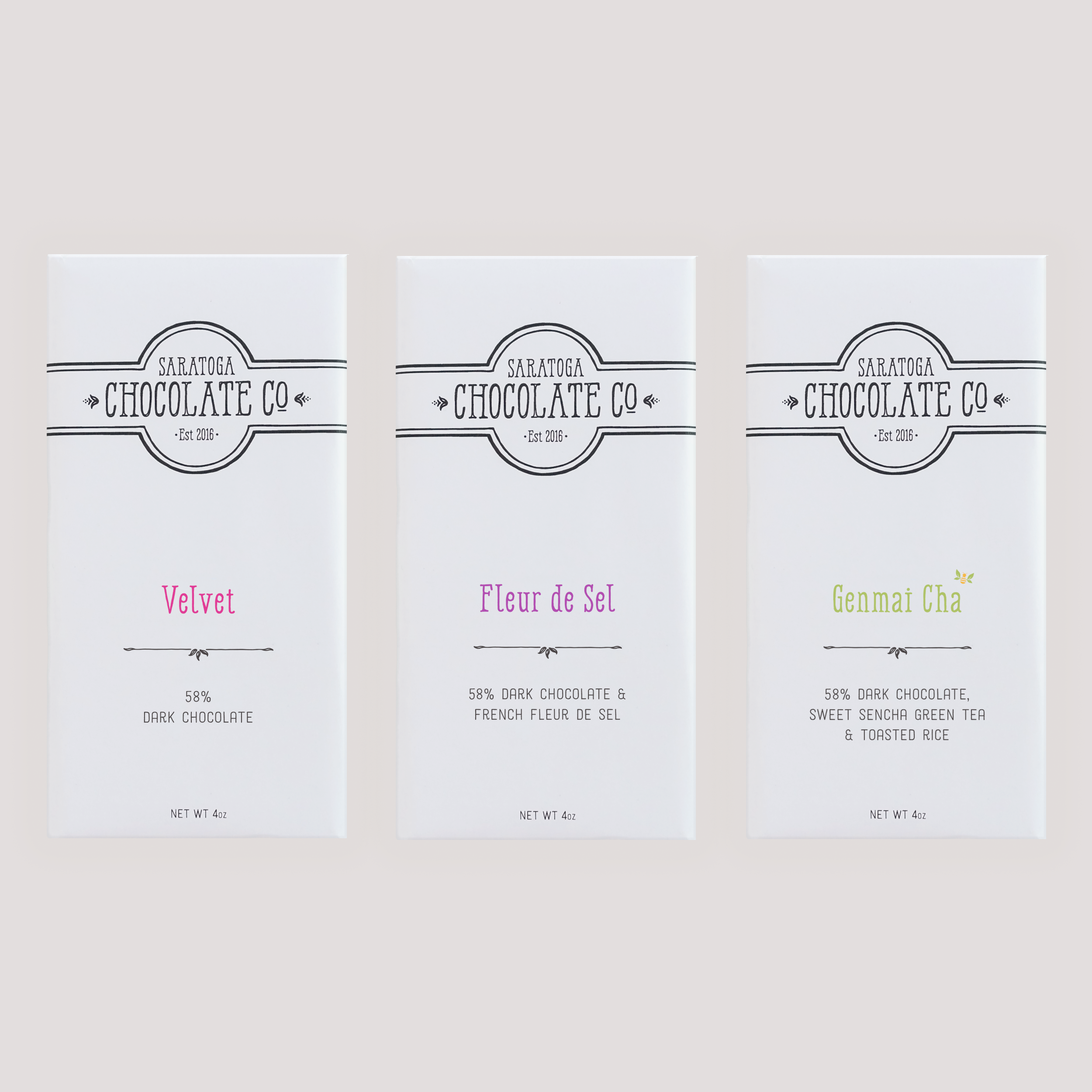 Three Saratoga Chocolate Co. dark chocolate bars in white wrappers with colorful text: Velvet, Fleur de Sel and Genmai Cha, displayed side by side on a light background.