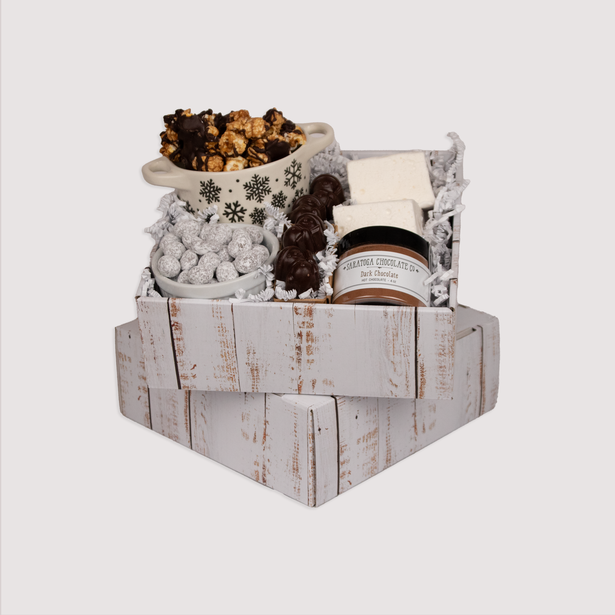 This distressed white wood style cardboard box is approximately 9 inches by 9 inches and three inches deep. Each box is filled with decorative white shredded paper and stuffed with a mini jar of dark chocolate hot chocolate, two freshly made pillow-soft, snow white vanilla marshmallows, four little dark chocolate caramel filled snowmen bonbons, powdered sugar dusted toffee chocolate covered pistachios, and a heaping serving of caramel corn with cacao nibs drizzled lightly in dark chocolate. 