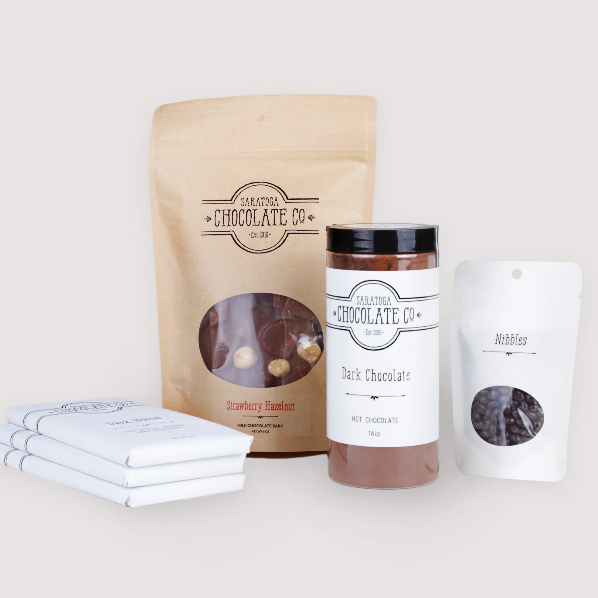 Gift set including three Saratoga Chocolate Co. bars, a Strawberry Hazelnut bark, a bag of dark chocolate nibbles, and a jar of dark chocolate hot chocolate, arranged together.