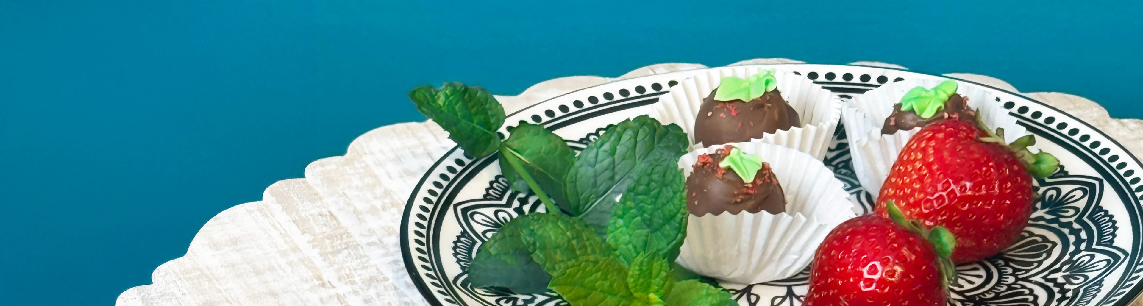 Dessert plate with chocolate truffles decorated with sugar leaf and strawberry topping, fresh strawberries and mint leaves. All displayed in a wooden tray and blue background