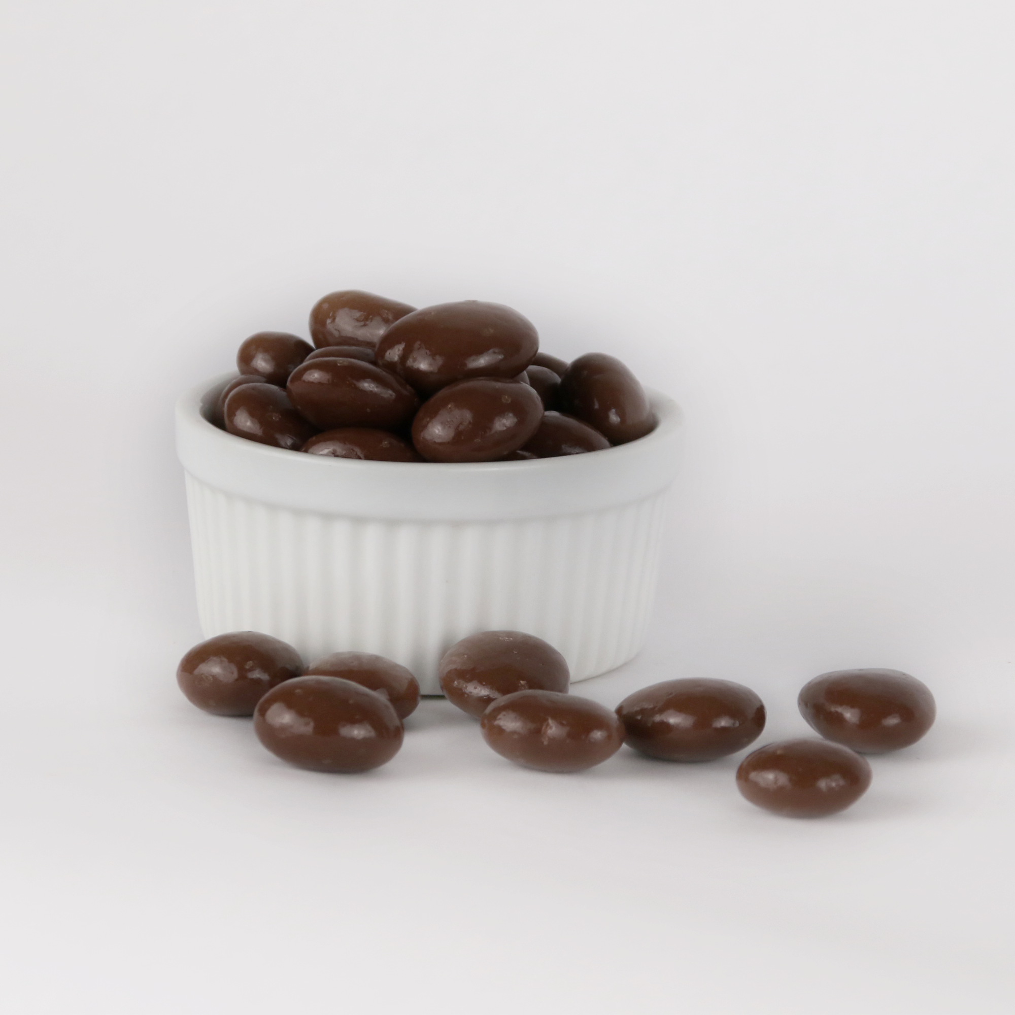 Image shows a pile of milk chocolate covered maple coated pecans displayed in a fluted white bowl with some milk chocolate covered maple coated pecans scattered in front of the bowl.