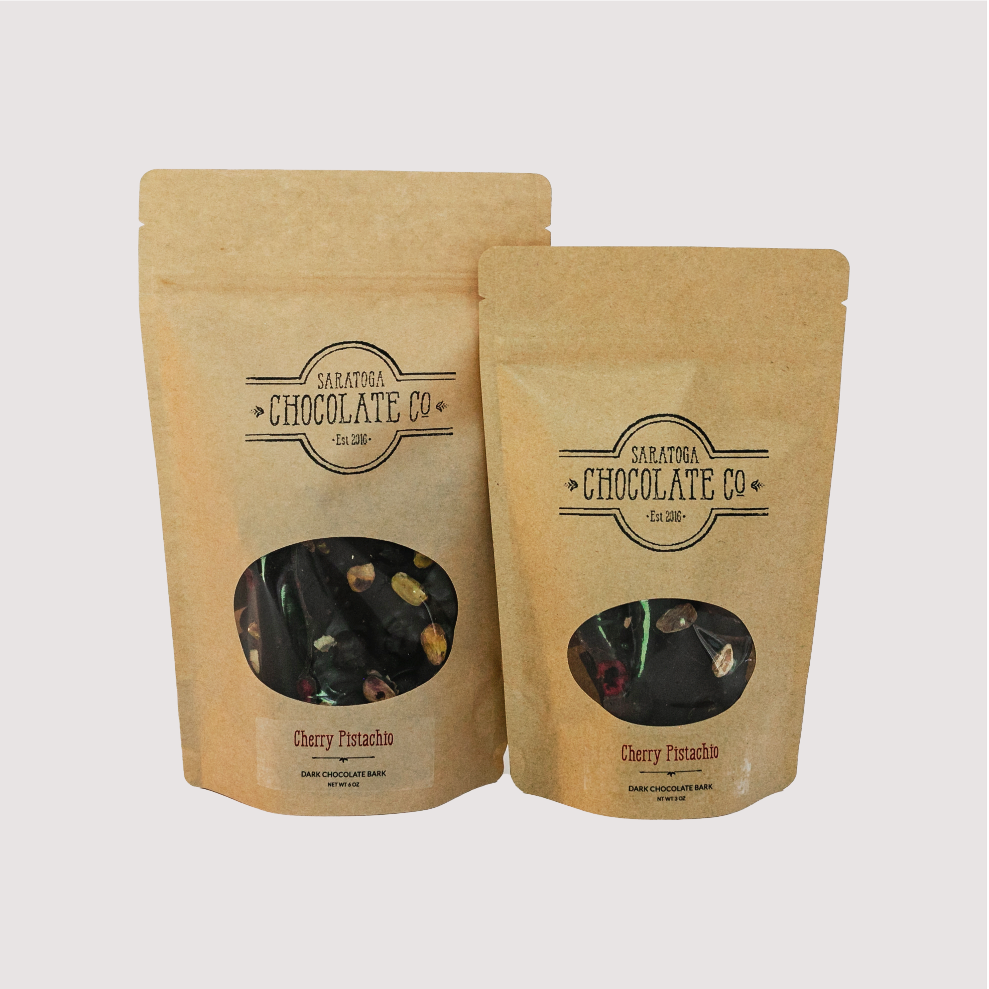 Saratoga Chocolate Co. Cherry Pistachio Dark Chocolate Bark packaged in kraft brown paper zipper sealed bags with a clear viewing window in 6-ounce and 3-ounce sizes, displaying the bark’s cherry and pistachio inclusions through the front of the bag. Each bag is sealed and labeled with the Saratoga Chocolate Co. logo and product name. This bark is vegan, gluten and dairy free.